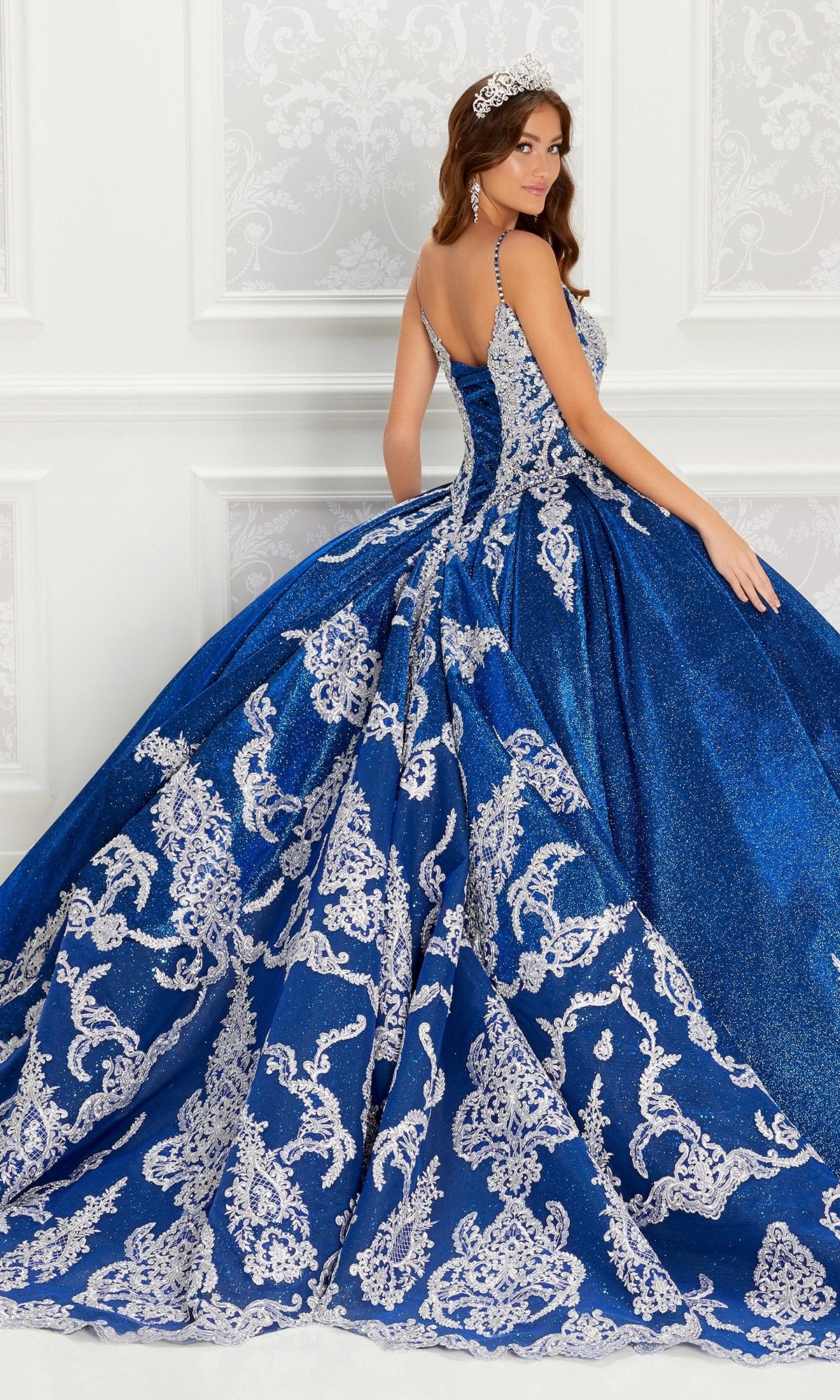 A woman wearing the Quinceanera Dress PR22141 by Princesa, featuring blue and silver embellishments, a corset back, full skirt, and train, stands in an elegant white-decorated room, looking over her shoulder.
